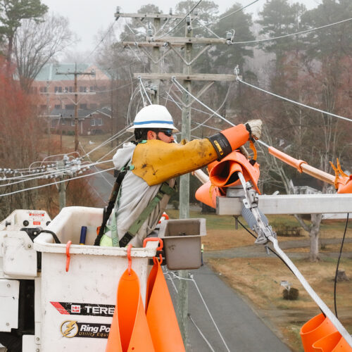 Utility worker in bucket truck covers power lines with orange sleeves.
