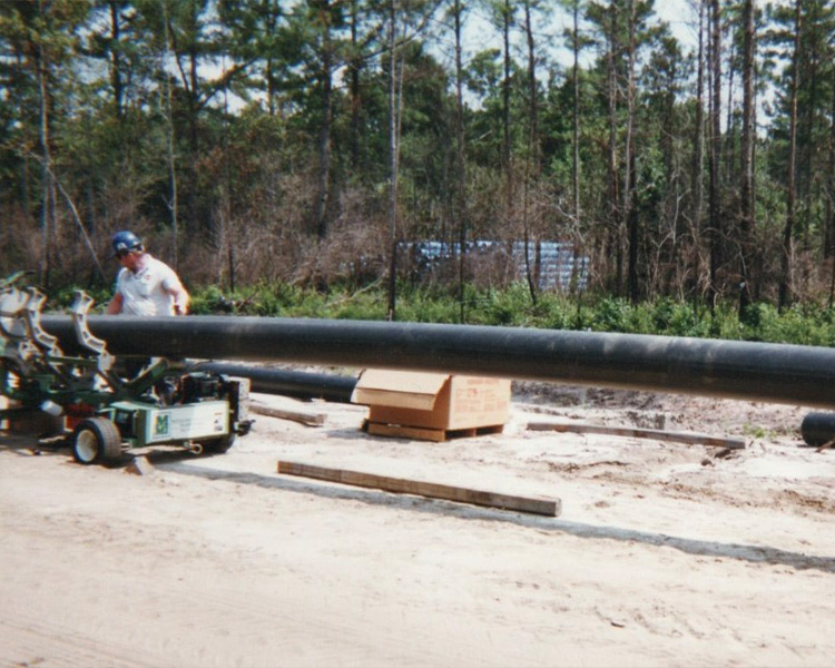 Worker joins large black pipes with machinery outdoors.