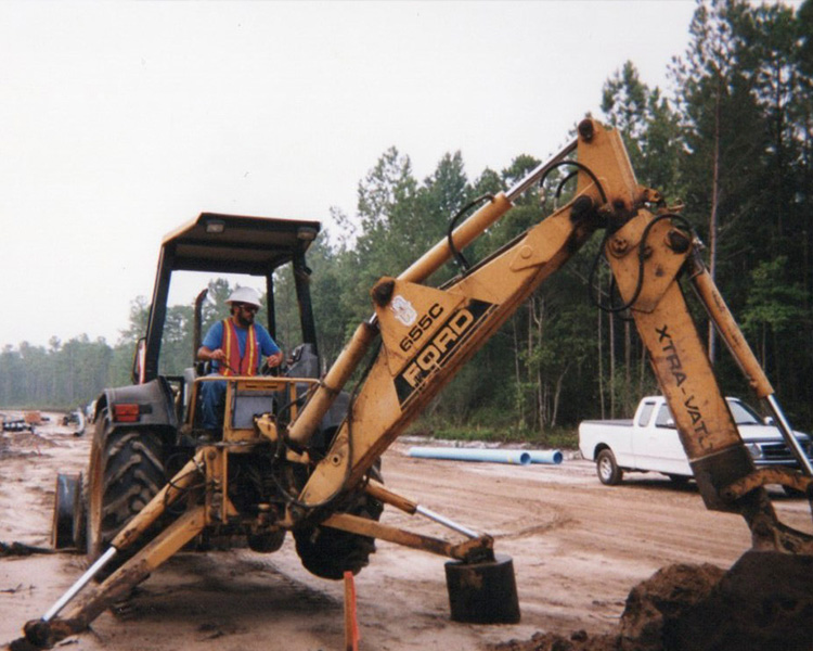 Worker drives Ford 655C backhoe loader on dirt road.
