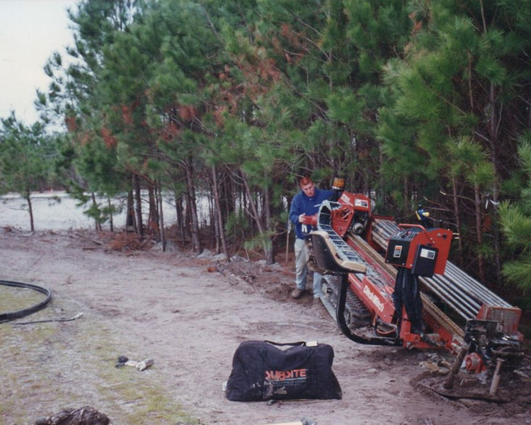 Person uses red drilling machine by trees; black bag nearby.