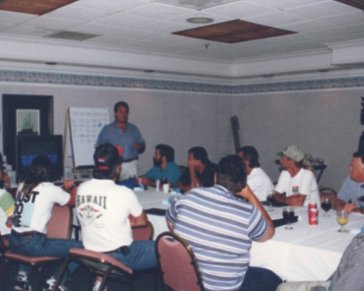 People sit at a table listening to someone speaking by a whiteboard.