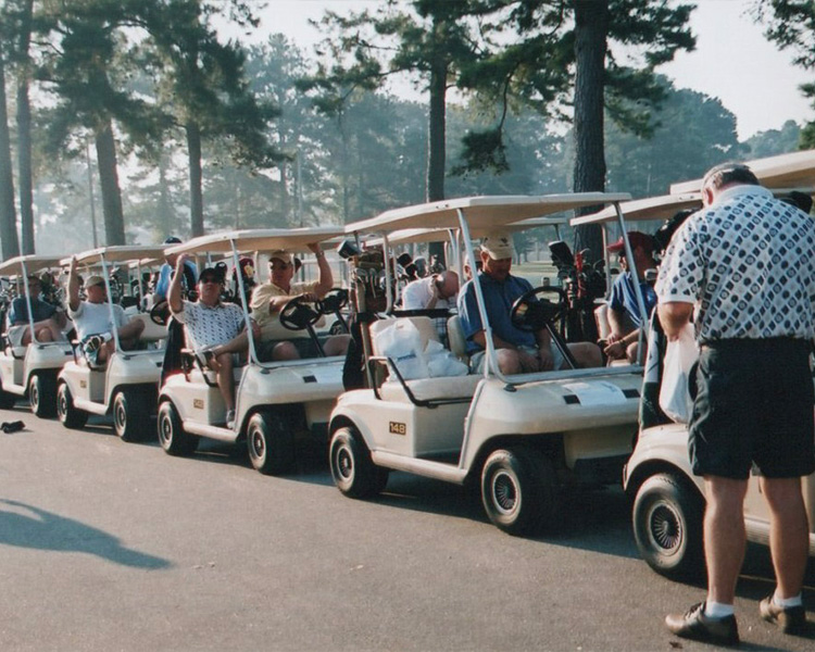 People sit in golf carts on a path; trees behind.