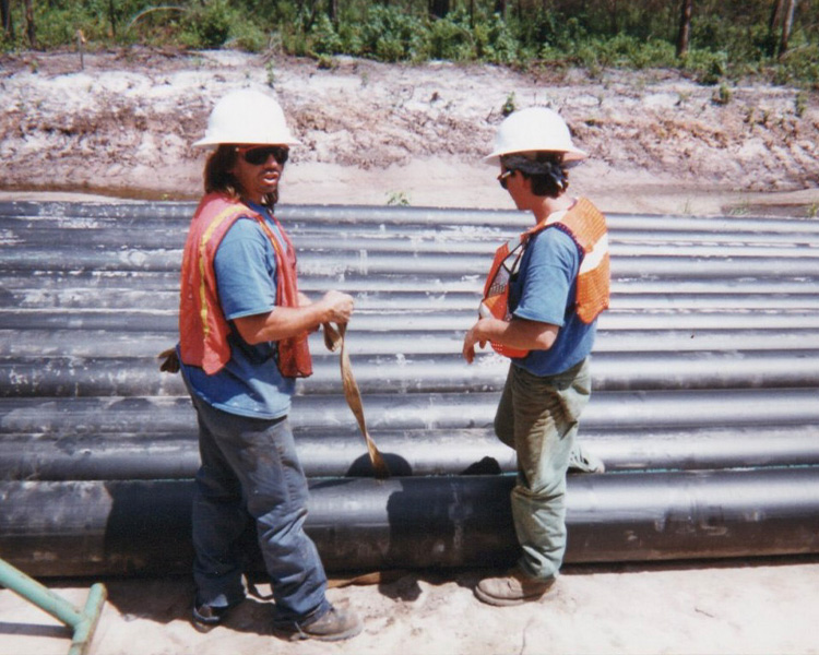 Two workers in safety gear stand by large black pipes outdoors.