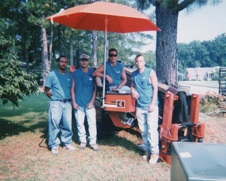 Four men in blue shirts stand under an orange umbrella by equipment.