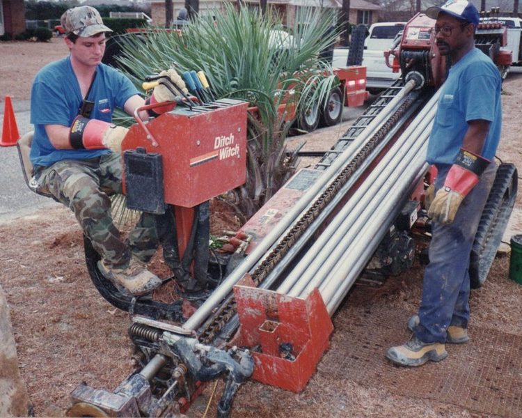 Two workers use a Ditch Witch drilling machine outside.