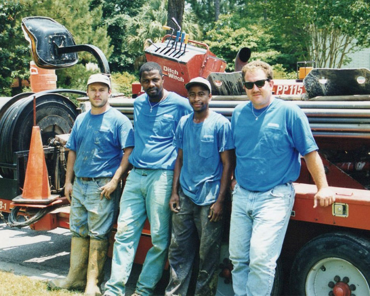 Four men in blue shirts by construction equipment and pipes.