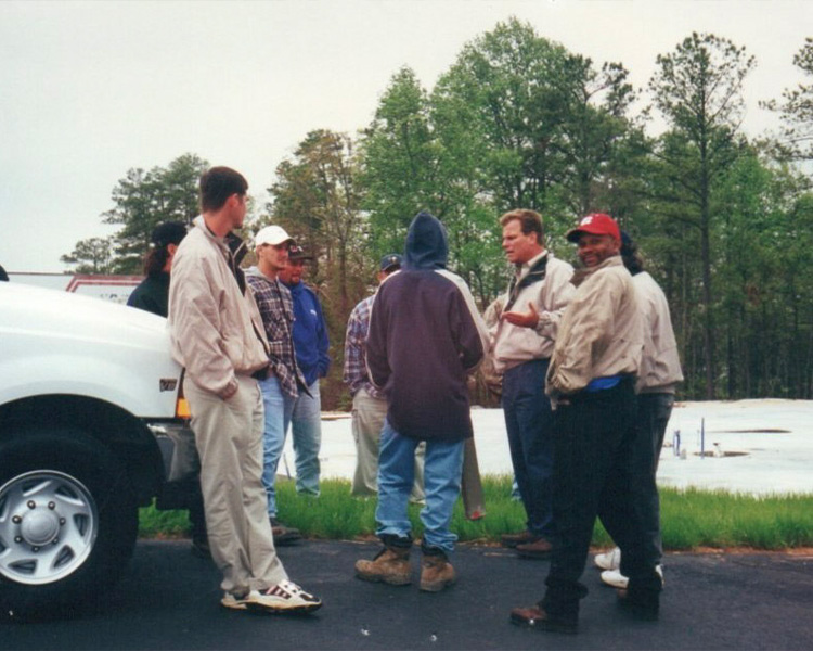 People talk by a white pickup truck in a tree-lined lot.