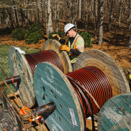 Worker in safety gear moves large orange cable reels outdoors.