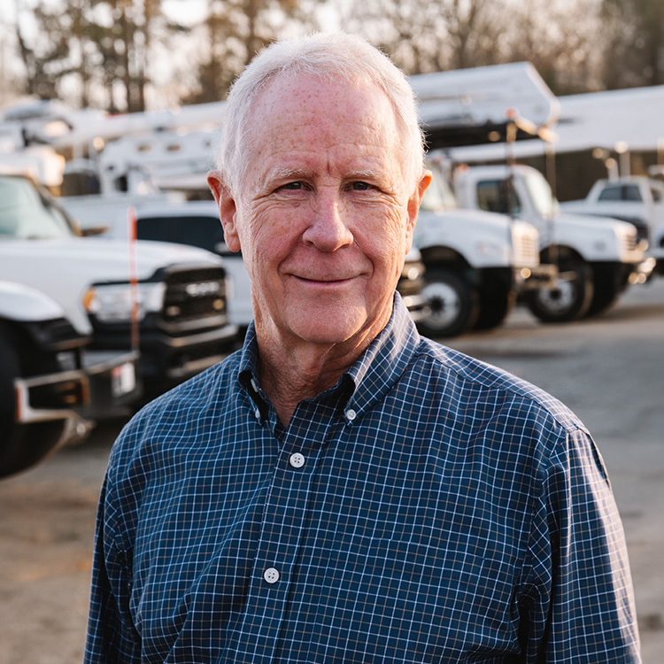 Older man in blue plaid shirt stands before white utility trucks.
