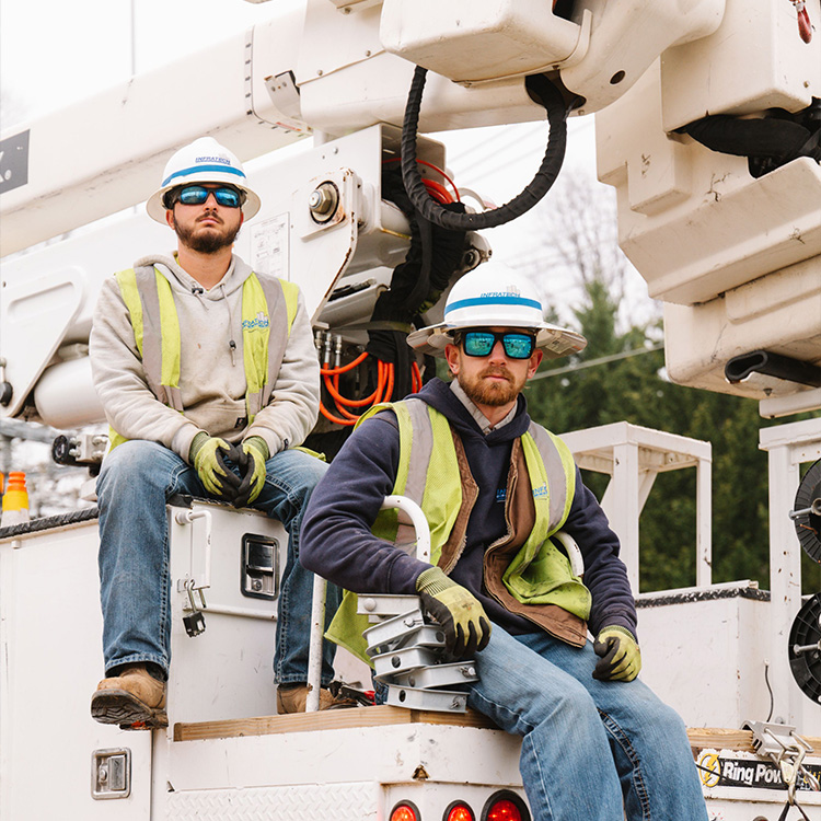 Two utility workers in safety gear sit on an equipment truck.