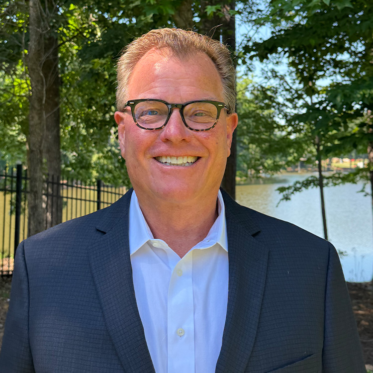 Smiling man in glasses and suit outdoors by water and trees.