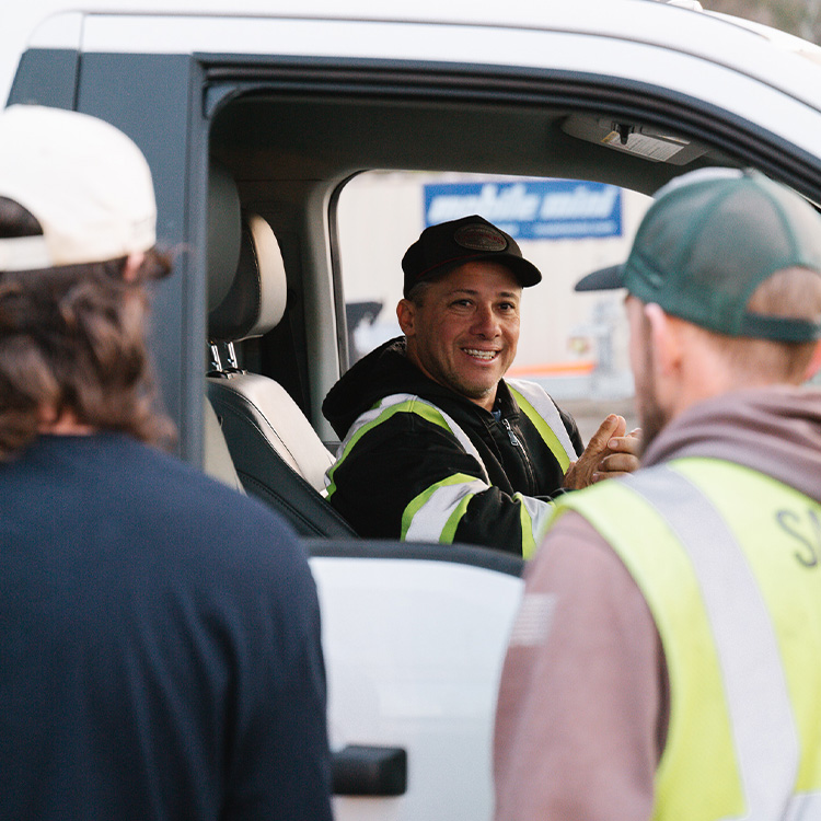 Smiling man in safety vest talks with two workers outside vehicle.
