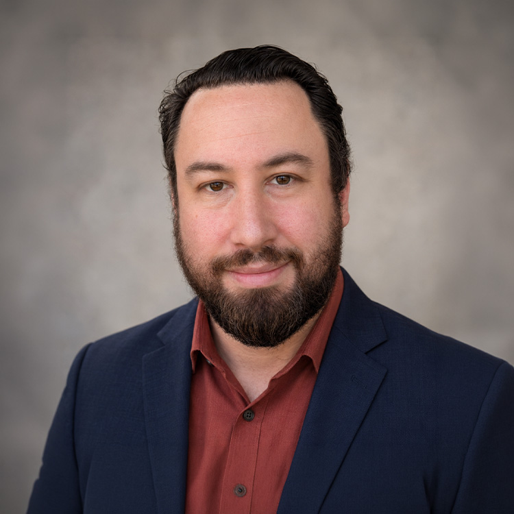 Man with dark hair, beard, navy jacket, rust shirt, gray background.