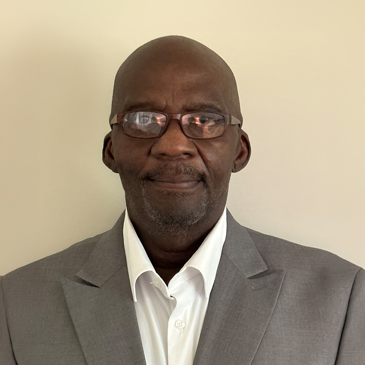 Man in glasses and grey suit jacket stands by plain background.