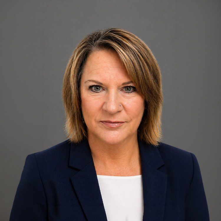 Middle-aged woman with light brown hair in navy blazer, gray background.