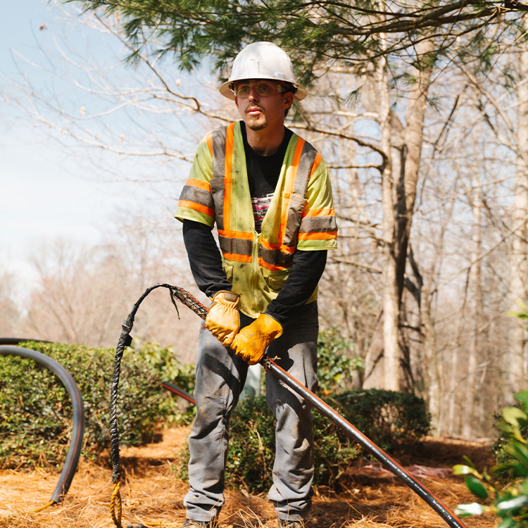 Construction worker in hard hat holds cable among trees.