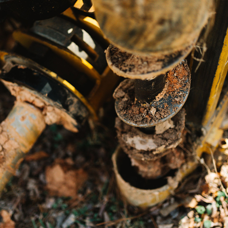 Dirty soil auger drill bit attached to equipment, ground visible.