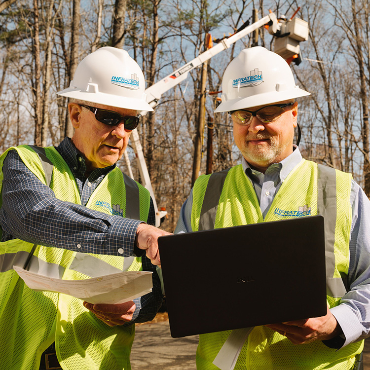 Men in safety vests review documents and laptop outdoors.