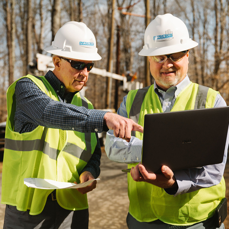 Two construction workers in safety gear review info on laptop.