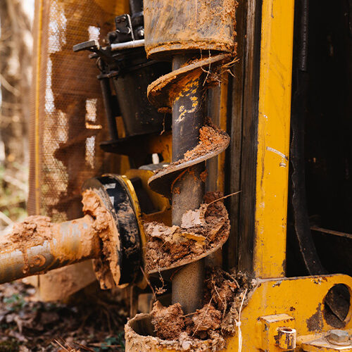 Muddy auger drill on yellow construction equipment with debris.